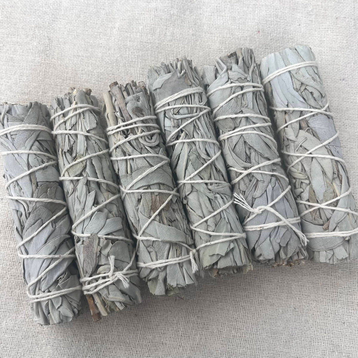 Five bundles of dried white sage smudge sticks tied with white string, arranged diagonally on a textured beige linen background.