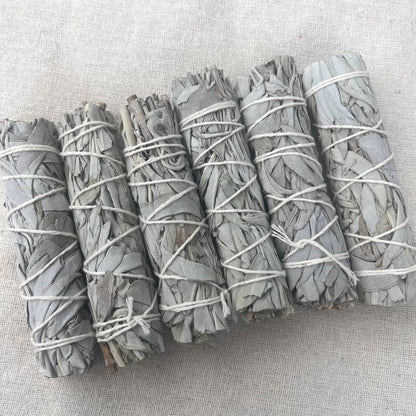 Five bundles of dried white sage smudge sticks tied with white string, arranged diagonally on a textured beige linen background.